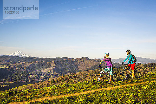 Zwei junge Frauen machen auf ihren Mountainbikes eine Pause  während sie auf einem einspurigen Weg durch eine offene Wiese mit Fluss und Vulkan in der Ferne fahren.