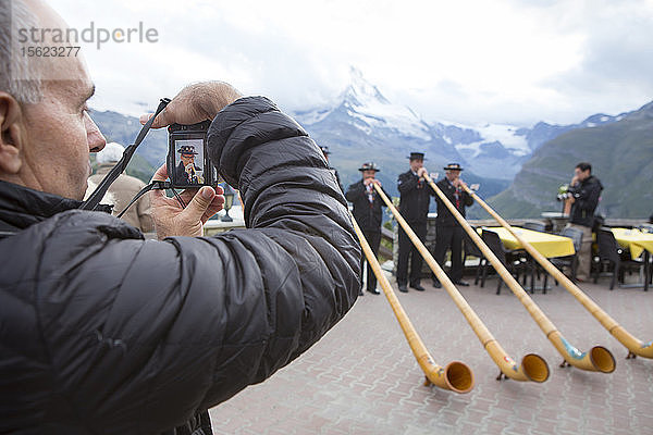 Ein Tourist fotografiert vier traditionell gekleidete Einwohner von Zermatt  die vor dem Matterhorn das Alphorn spielen. Im Laufe der Zeit verschwand das Alphorn als Instrument der Schweizer Hirten fast völlig. Erst mit der Romantik des 19. Jahrhunderts und der Wiederbelebung von Folklore und Tourismus erlebte das Alphorn eine Renaissance und wurde sogar zu einem nationalen Symbol.