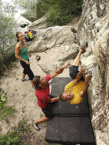 Zwei Männer bouldern im Valle Maggia  einem Bergtal in der Nähe von Locarno in der Schweizer Provinz Tessin. Die Felsen in dieser Region  die für ihr warmes Wetter bekannt ist  bieten hervorragende Klettermöglichkeiten. Ein Kletterer versucht  ein Boulderproblem zu senden  der andere weist auf einen guten Halt hin. Eine Kletterin entspannt sich  während sie darauf wartet  dass sie an der Reihe ist.