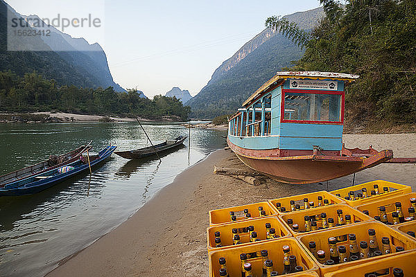 Boote und ein großer Stapel leerer Beer Lao (das nationale Bier) am Ufer des Nam Ou Flusses in Muang Ngoi  Laos. Das Bier ist der notwendige Treibstoff für den lebhaften Tourismus  der sich um die Stadt herum abspielt.
