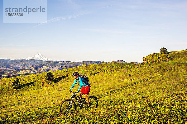 Eine junge Frau fährt mit dem Mountainbike auf einem einspurigen Weg durch grünes Gras in der frühen Morgensonne mit einem Vulkan in der Ferne.