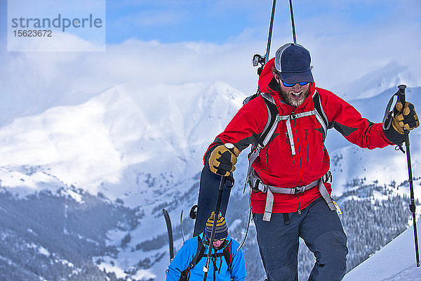 Ein Skifahrer und ein Snowboarder wandern auf einem Berg mit schneebedeckten Gipfeln in der Ferne.