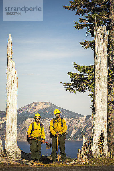 Zwei Feuerwehrleute in gelben Hemden stehen mit ihrer Ausrüstung zwischen zwei abgestorbenen Bäumen  im Hintergrund ein Bergsee. Crater Lake  Oregon  USA