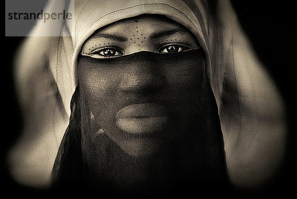 Porträt einer Frau mit verziertem Gesicht hinter transparentem schwarzem Schleier auf schwarzem Hintergrund  Meknes  Marokko