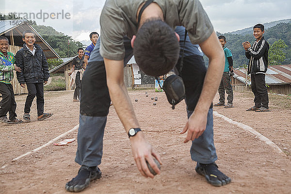 Robert Hahn spielt mit Schülern eines Internats Boule (p??tanque)  wobei er die Kugel scherzhaft zwischen seine Beine wirft  in Ban Tang  Laos.