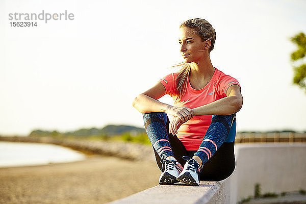 Eine junge sportliche Frau  die an der Wand sitzt und die Aussicht auf den Strand genießt