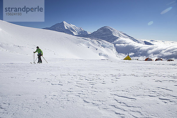 Ein Skibergsteiger auf dem Kahiltna-Gletscher im Denali-Nationalpark in Alaska  mit dem Berg Foraker und dem Kahiltna Dome im Hintergrund. Rechts sind die Zelte des 11 K Camps am Denali zu sehen.