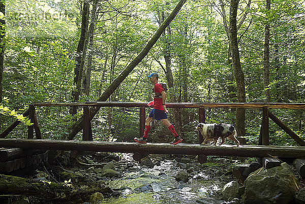 Mann mit seinem Hund Wandern auf Holzbrücke durch einen Bach im Wald übergeben