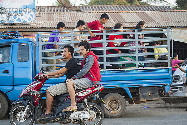 Junge Männer fahren gemeinsam auf einem Motorroller an einem Lastwagen vorbei  der auf einer belebten Straße in Luang Prabang  Laos  entladen wird.