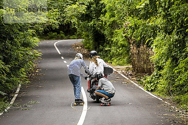 Zwei Longboarder tragen ein Motorrad hinter sich her  das einen dritten Skateboarder trägt