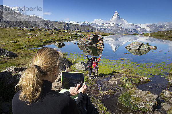 Ein Tourist macht mit einem Ipad-Tablet ein Foto von zwei anderen Personen vor dem Stellisee  in dem sich das Matterhorn spiegelt. Dieser Bergsee oberhalb von Zermatt in den Schweizer Alpen ist ein beliebtes Touristenziel.