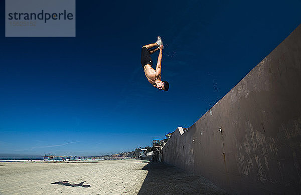 Ein athletischer Mann vollführt einen Rückwärtssalto von einer Zementwand auf den Strand in San Diego  Kalifornien.