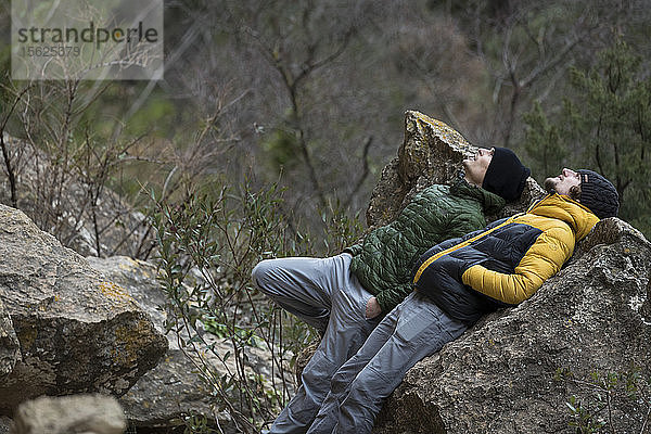 Seitenansicht von zwei Männern in Jacken  die auf dem Rücken an einem Felsen liegen und nach oben schauen