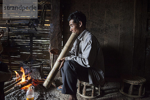 Ein Mann raucht in einem Haus in Ban Sop Kha  Laos  eine Zigarette durch eine große Bambus-Wasserpfeife am Feuer.