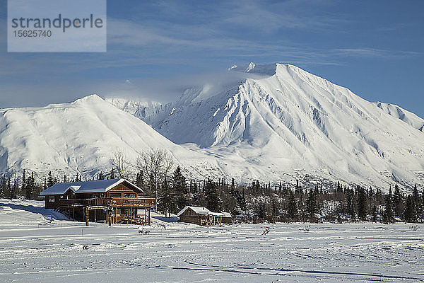 Blockhütten in Alaska liegen am Fuße eines zugefrorenen Sees mit schneebedeckten Bergen im Hintergrund.