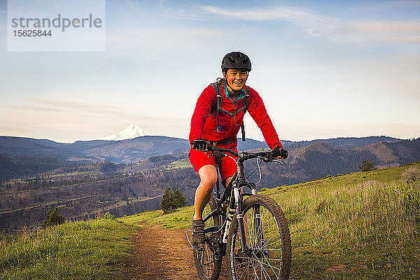 Eine junge Frau in einem roten Mantel fährt mit einem Mountainbike auf einem einspurigen Weg durch grünes Gras im frühen Morgenlicht.