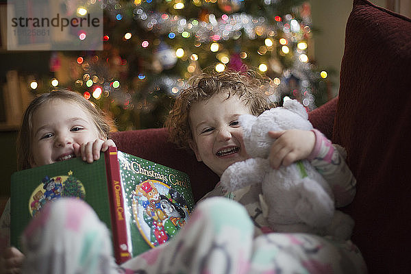 Lächelndes Mädchen mit Bilderbuch und Junge mit Teddybär auf dem Sofa sitzend  mit Weihnachtsbaum im Hintergrund