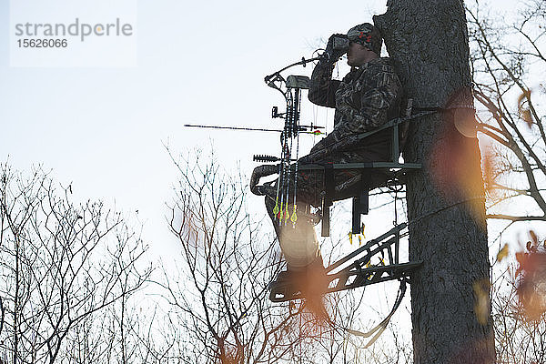 Junger erwachsener männlicher Jäger  der auf einem Baum sitzt und nach Rehen Ausschau hält
