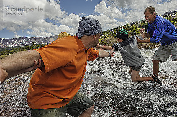 Zwei Männer helfen einem Jungen  der von Stein zu Stein über einen reißenden Bach  einen Nebenfluss des Yellowstone Creek  in der alpinen Tundra unterhalb des Kings Peak springt  am vierten Tag der sechstägigen Rucksacktour von Troop 693 in der High Uintas Wilderness Area  Uintas Range  Utah
