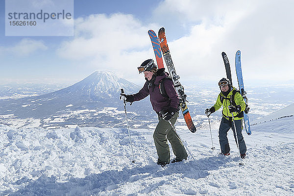 Mit dem Vulkan Yotei im Hintergrund wandern eine Skiläuferin und ein Skiläufer zum Gipfel des Annupuri in der Nähe des Skigebiets Niseko United auf der japanischen Insel Hokkaido. Die Skier  die sie in ihrem Rucksack tragen  sind groß und breit  so dass die Wintersportler die hüfttiefen Tiefschneehänge  für die Niseko United bekannt ist  in vollen Zügen genießen können. Niseko United besteht aus vier Skigebieten auf einem Berg  dem Annupuri (1.308 m). Der 100 km südlich von Sapporo gelegene Niseko Annupuri ist ein Teil des Quasi-Nationalparks Niseko-Shakotan-Otaru Kaigan und der östlichste Park der Niseko-Vulkangruppe. Hokkaido  die nördliche Insel Japans  liegt geografisch ideal in der Bahn der beständigen Wettersysteme  die die kalte Luft aus Sibirien über das Japanische Meer bringen. Dies führt dazu  dass viele der Skigebiete mit Pulverschnee überhäuft werden  der für seine unglaubliche Trockenheit bekannt ist. In einigen der Skigebiete Hokkaidos fallen durchschnittlich 14-18 Meter Schnee pro Jahr. Niseko ist die Pulverschneehauptstadt der Welt und als solche das beliebteste internationale Skigebiet in Japan. Es bietet ein unvergessliches Erlebnis für Skifahrer und Snowboarder aller Niveaus. Der Berg Yotei im Hintergrund wird oft auch als der Berg Fuji von Hokkaido bezeichnet.