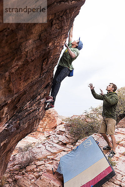 Ein sportlicher Mann bouldert auf einem hohen Felsen  während ein anderer Mann ihn von unten in Red Rocks  Nevada  entdeckt.