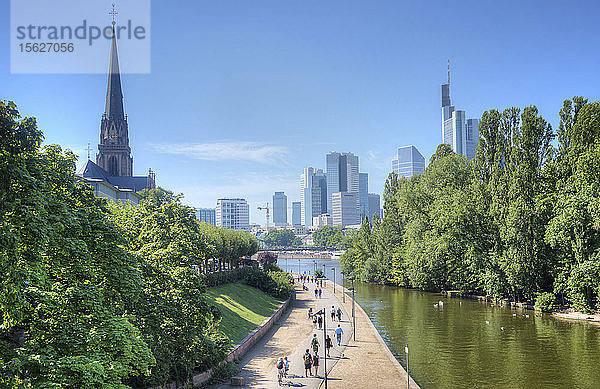 Besucher schlendern an einem schönen Tag entlang der Mainpromenade mit der Frankfurter Skyline und hohen Bäumen im Hintergrund.