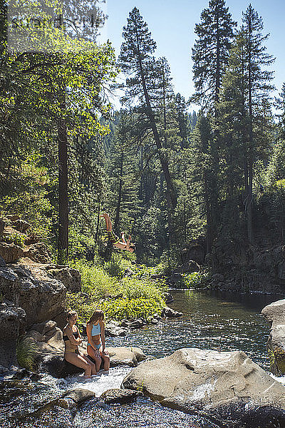 Blick auf zwei junge Frauen  die auf einem Felsen im Fluss sitzen  und einen Mann  der von einer Klippe springt  McCloud River  Kalifornien  USA