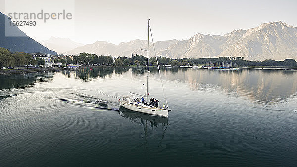 Luftaufnahme eines Segelbootes und eines Rettungsbootes  die den Genfer See überqueren  mit Bergen im Hintergrund  Bäumen am Seeufer und einem Jachthafen in Villeneuve  Kanton Waadt  Schweiz