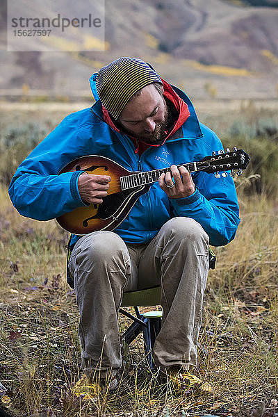 Männlicher Camper in Jacke und Strickmütze  der sitzend Mandoline spielt  Jackson  Wyoming  USA