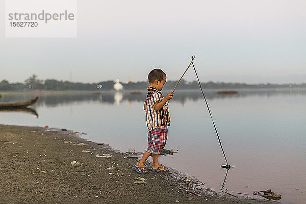 Kleiner Junge steht am Seeufer und spielt mit einer selbstgebauten Angelrute  Mandalay  Mandalay District  Myanmar