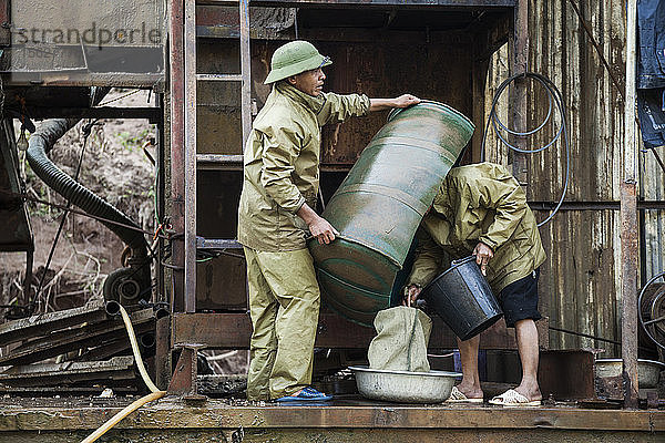 Vietnamesische Männer leeren das Konzentrat aus dem Schleusenkasten in einen Sack für die spätere Verarbeitung auf dem Deck eines mechanischen Goldbaggers  der auf dem Nam Ou Fluss in Laos schwimmt.