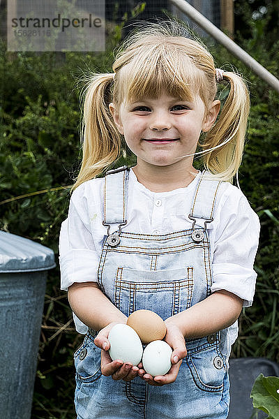 Blondes Mädchen steht in einem Garten  hält frische Eier in der Hand und lächelt in die Kamera.