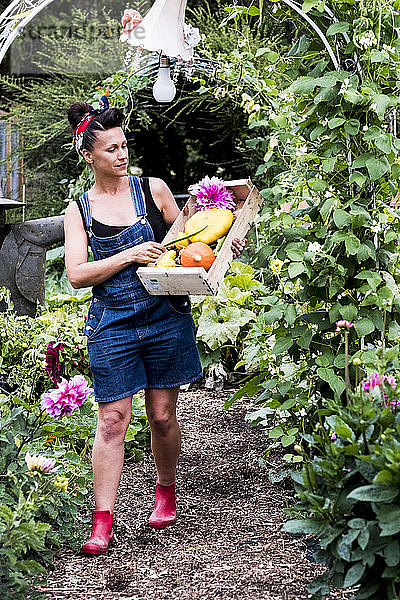 Frau  die in einem Garten spazieren geht und eine Holzkiste mit frischem Gemüse und Blumen trägt.