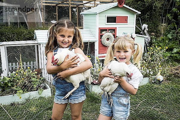 Zwei Mädchen stehen vor einem Hühnerstall in einem Garten  halten weiße Hühner und lächeln in die Kamera.