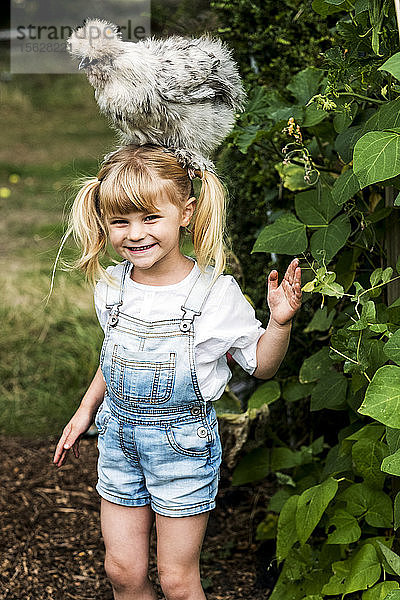 Lächelndes blondes Mädchen  das in einem Garten steht  mit einem flauschigen grauen Huhn auf dem Kopf.