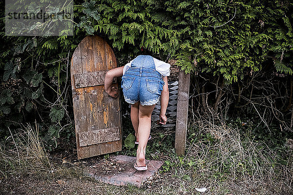 Rückansicht eines Mädchens in Denim-Latzhose  das durch eine kleine Holztür in einer Gartenhecke geht.