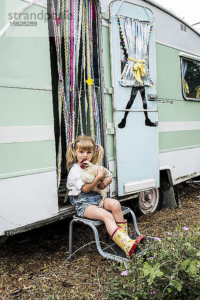 Blondes Mädchen hält weißes Huhn  das vor einem Wohnwagen sitzt.