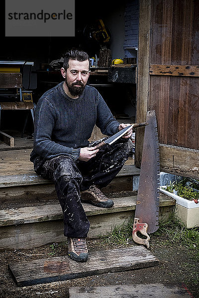 Bärtiger Mann sitzt auf Stufen vor seiner Werkstatt  hält ein handgefertigtes Messer in der Hand und schaut in die Kamera.