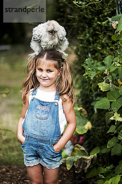 Lächelndes blondes Mädchen  das in einem Garten steht  mit einem flauschigen grauen Huhn auf dem Kopf.