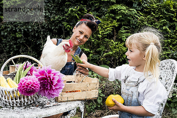 Frau und Mädchen sitzen an einem Tisch in einem Garten und füttern weiße Hühner.