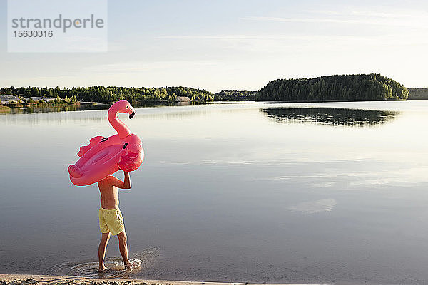 Junger Mann mit Flamingopool schwimmt am Seeufer spazieren