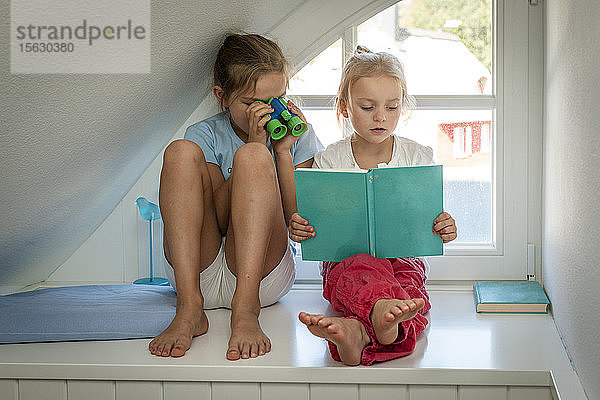 Mädchen mit Spielzeugfernglas schaut Schwester mit Buch am Fenster an