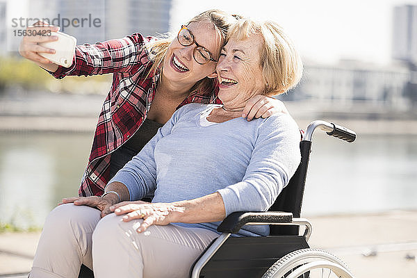 Junge Frau mit ihrer lächelnden Großmutter im Rollstuhl sitzend und mit einem Selfie