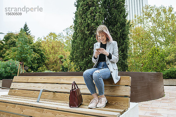 Lächelnde Frau sitzt auf Parkbank und benutzt Mobiltelefon
