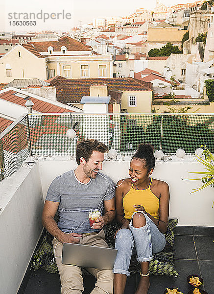 Glückliches junges Paar mit Laptop und Kopfhörern sitzt abends auf dem Dach  Lissabon  Portugal