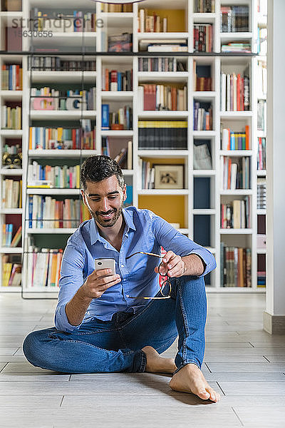 Porträt eines lächelnden jungen Mannes  der zu Hause vor Bücherregalen auf dem Boden sitzt und auf ein Smartphone schaut