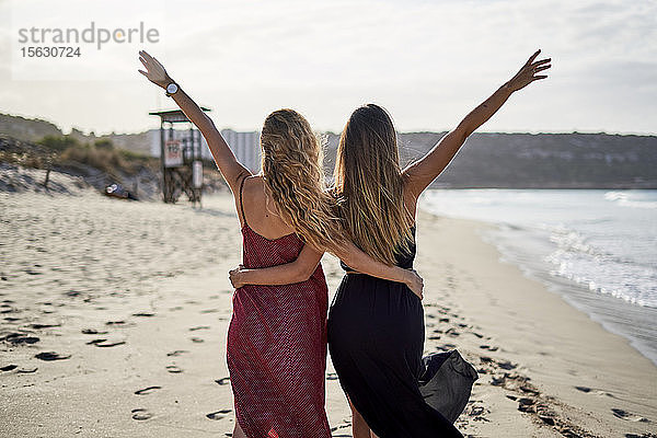 Zwei junge Frauen stehen Arm in Arm mit ausgestreckten Armen an einem Strand