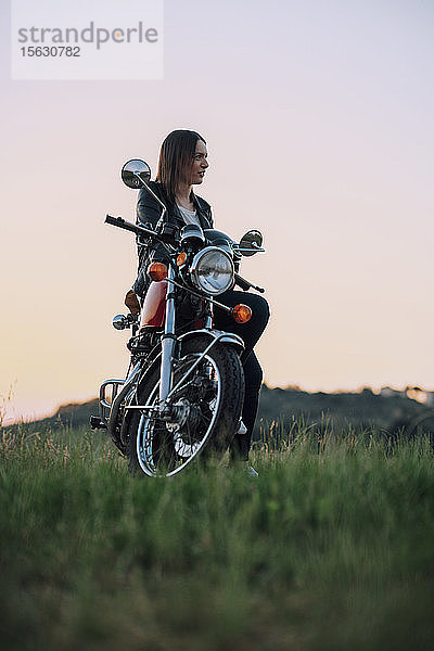 Junge Frau mit Oldtimer-Motorrad in ländlicher Szene genießt Sonnenuntergang