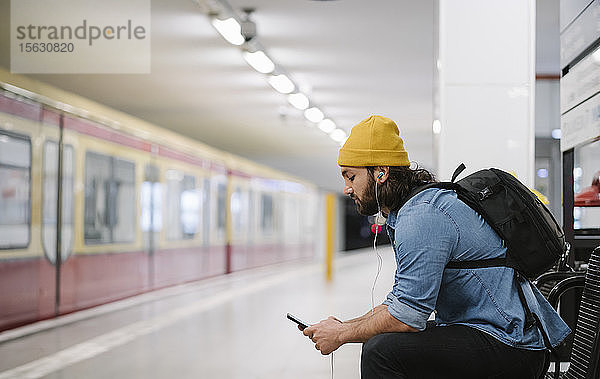 Mann mit Rucksack hört mit Smartphone und Kopfhörern Musik  während er am Bahnsteig wartet  Berlin  Deutschland
