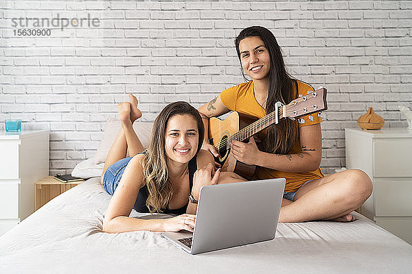 Porträt eines glücklichen lesbischen Paares mit Laptop und Gitarre im Bett